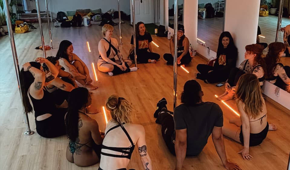 Dancers sitting in a circle on the studio floor between the poles, talking and laughing